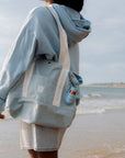 Person wearing a light blue hoodie and shorts with a matching bag on a beach.