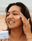 Smiling woman applying Bloomeffects Tulip Screen Whipped Butter Crème with SPF 50 to her cheek on the beach.