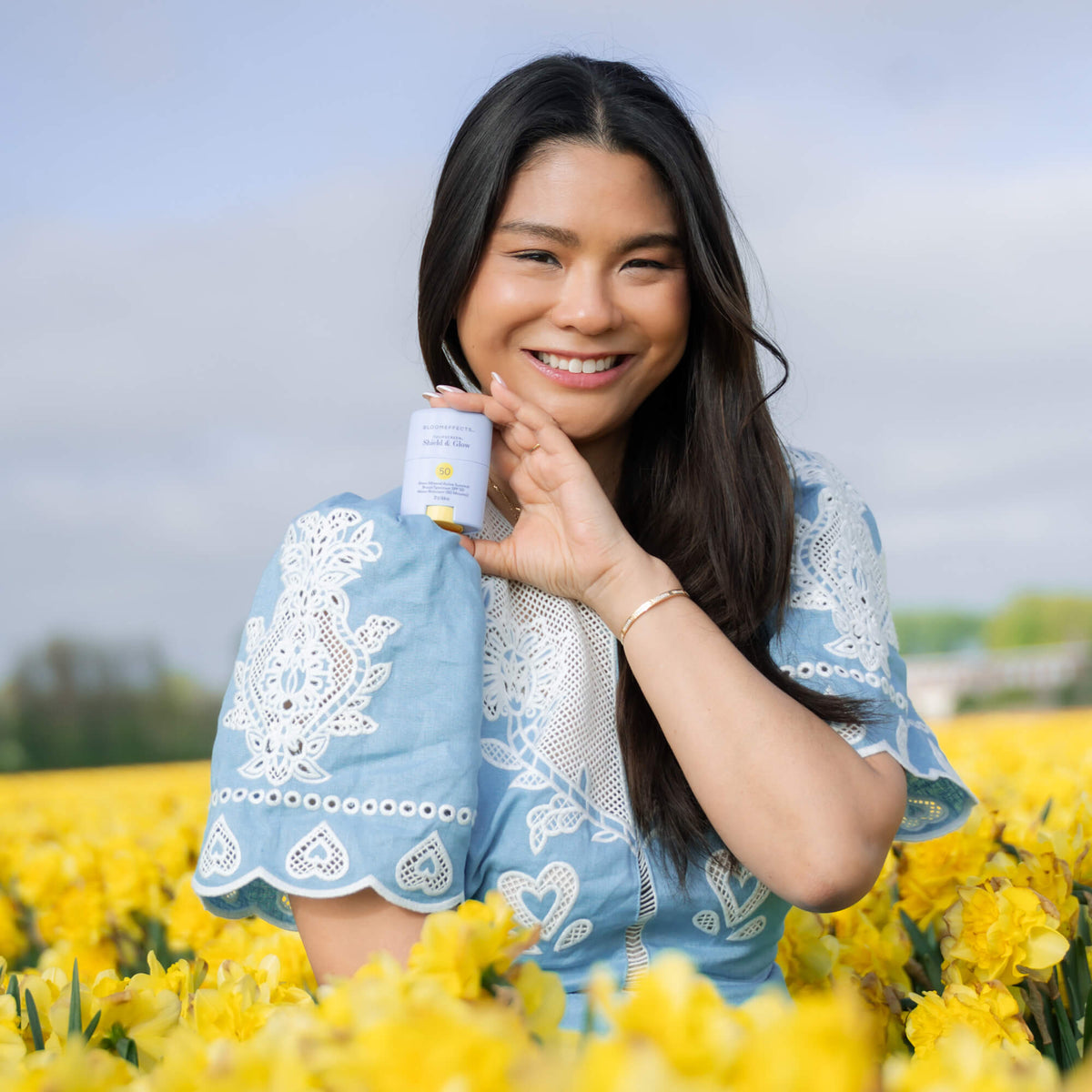 Smiling woman in a blue embroidered dress holding Bloomeffects Tulipscreen Shield &amp; Glow SPF 50 sunstick in a field of yellow tulips
