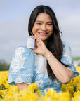 Smiling woman in a blue embroidered dress holding Bloomeffects Tulipscreen Shield & Glow SPF 50 sunstick in a field of yellow tulips