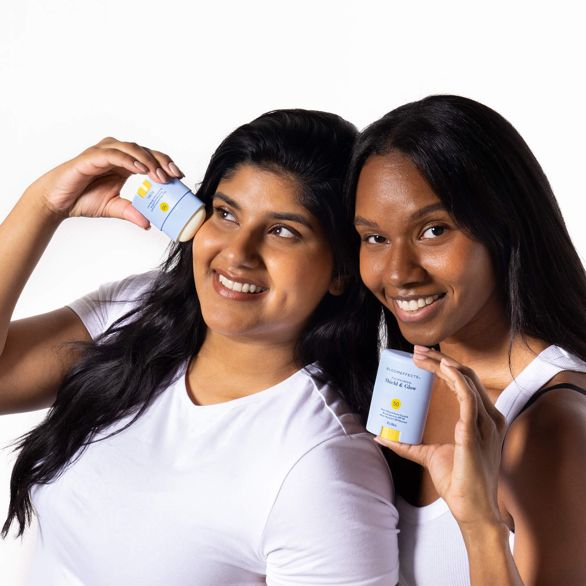 Two smiling women in white tops, one applying Bloomeffects Tulipscreen Shield &amp; Glow sunstick to her cheek and the other holding the stick beside her face