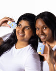 Two smiling women in white tops, one applying Bloomeffects Tulipscreen Shield & Glow sunstick to her cheek and the other holding the stick beside her face