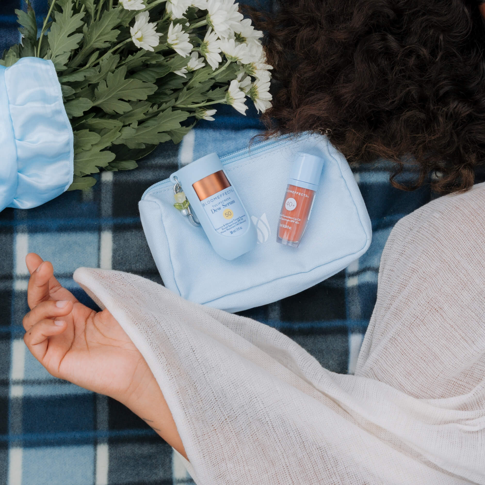 Cosmetic bag with skincare products on a checkered blanket, with a person's hand and flowers in the background.