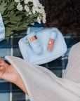 Cosmetic bag with skincare products on a checkered blanket, with a person's hand and flowers in the background.