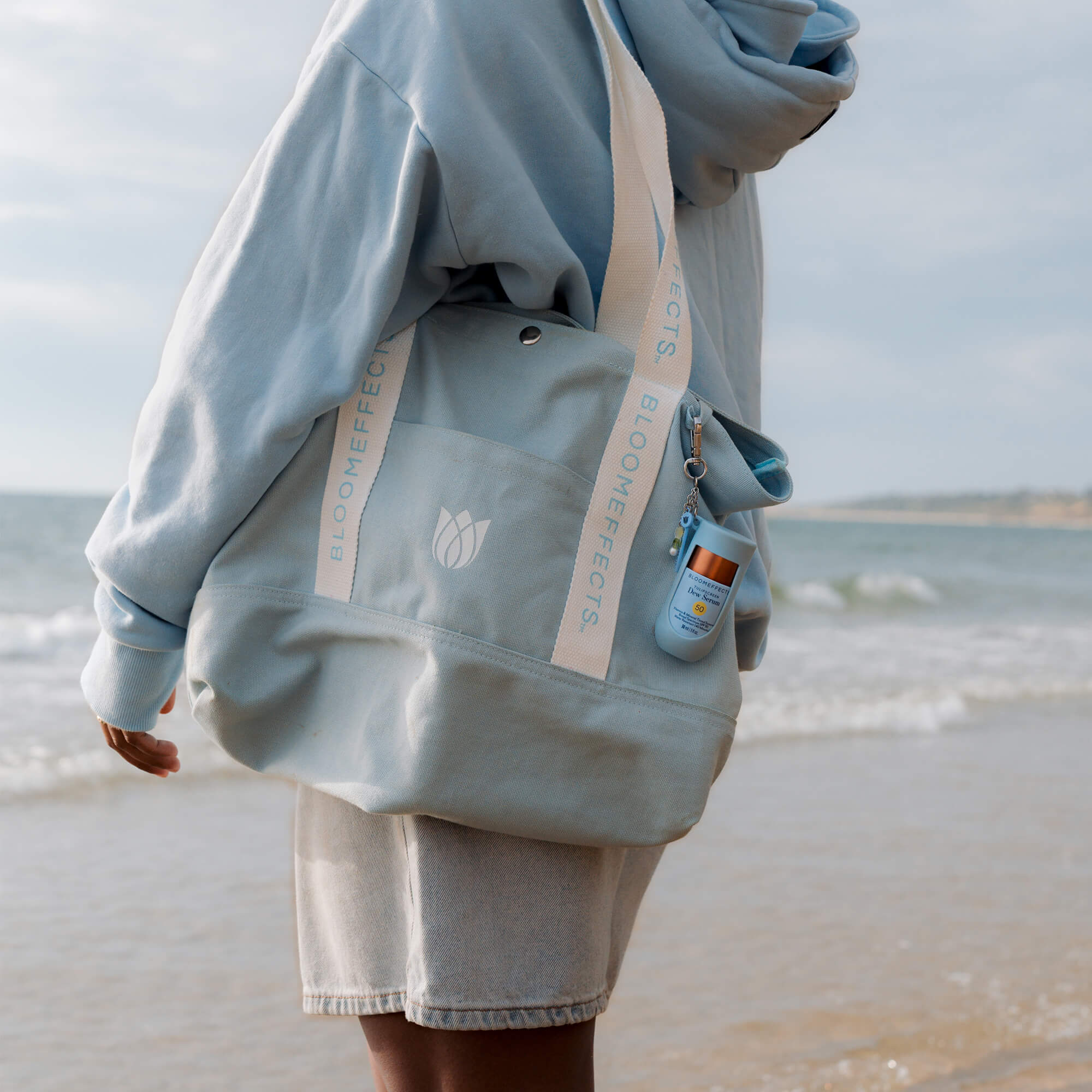 Person wearing a light blue hoodie and shorts with a Bloomeffects bag on a beach.