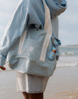 Person wearing a light blue hoodie and shorts with a Bloomeffects bag on a beach.