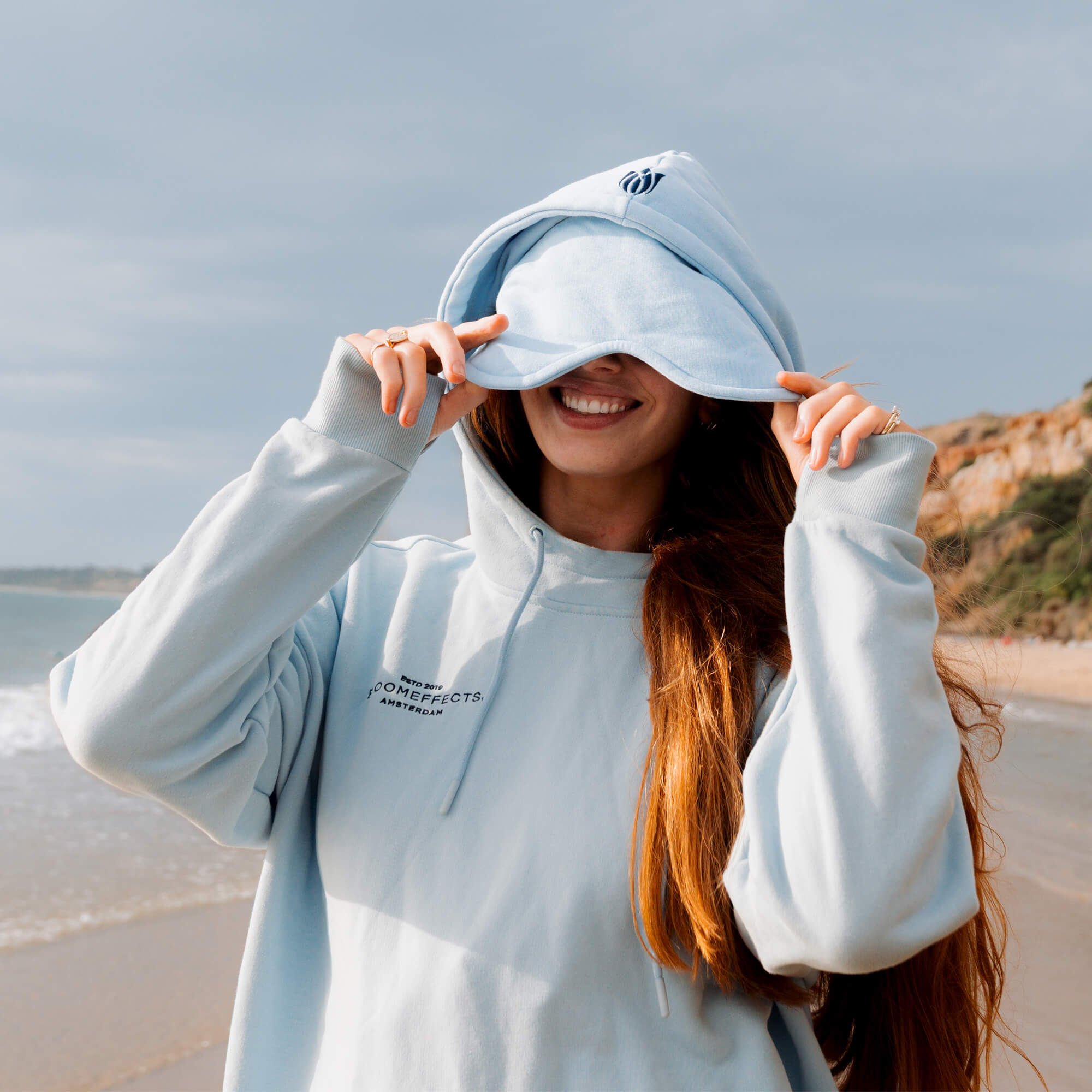 Person wearing a light gray hoodie with a logo on a beach