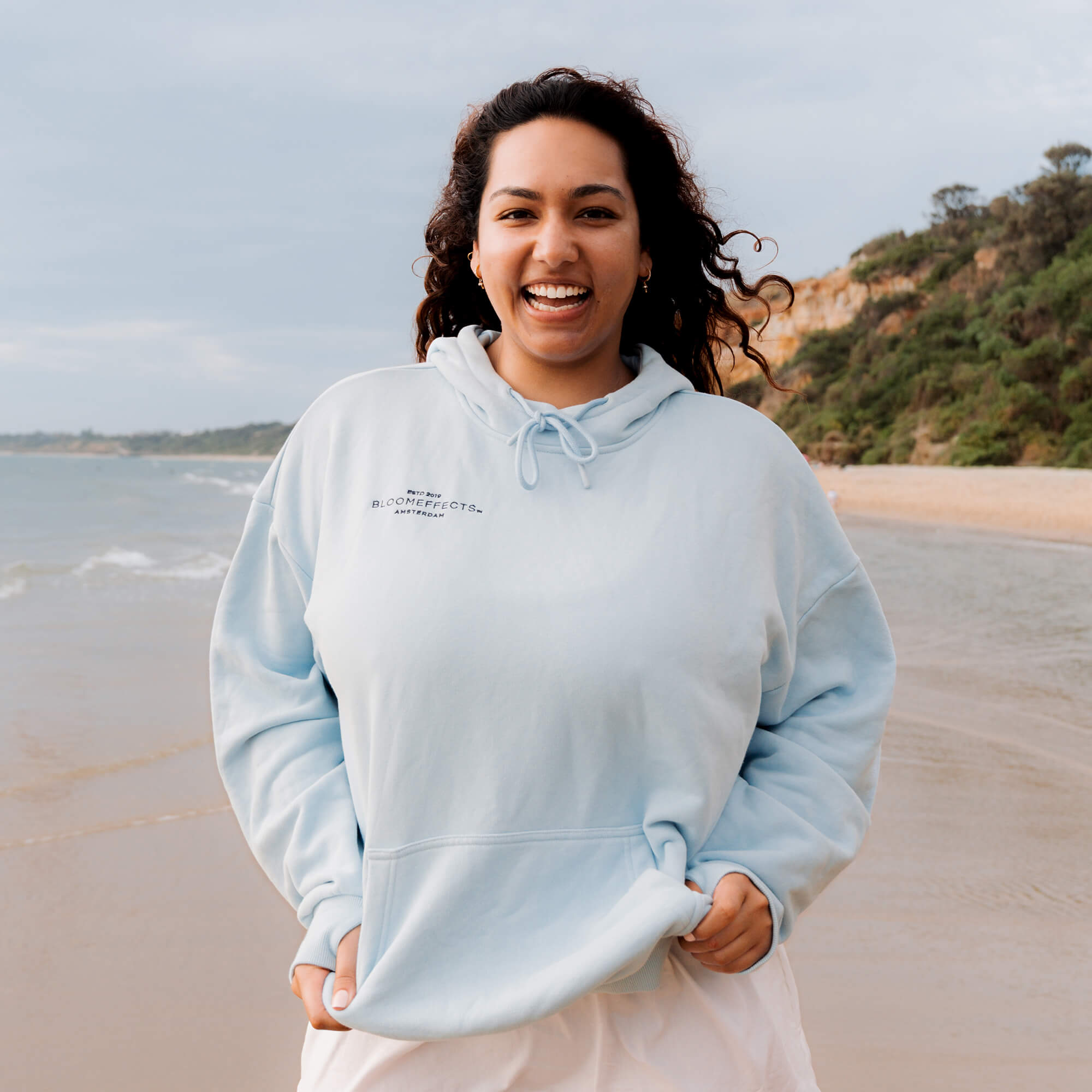Woman wearing a light blue hoodie on a beach with ocean and cliffs in the background