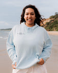 Woman wearing a light blue hoodie on a beach with ocean and cliffs in the background