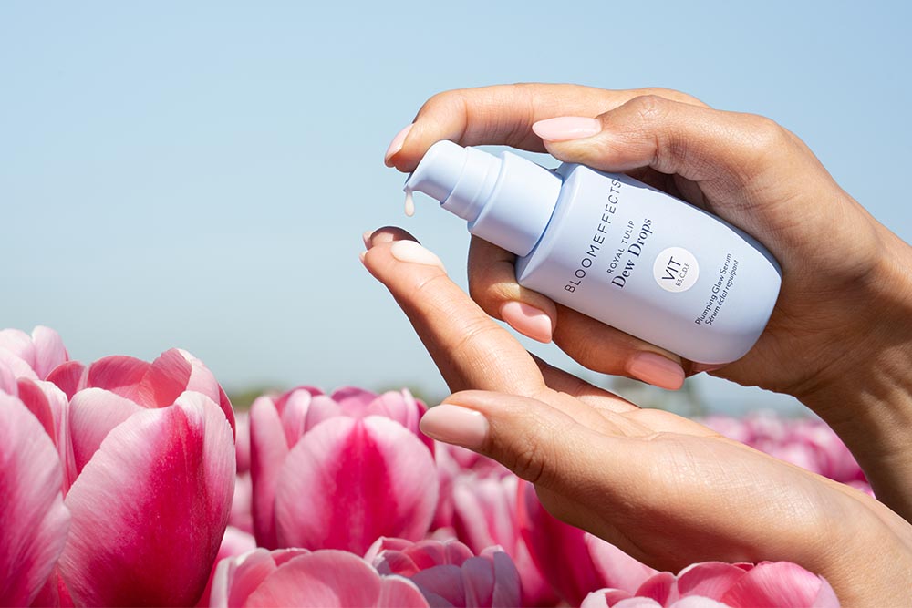 A person's hands holding and pumping a bottle of Bloomeffects Dew Drops set against a background of pink tulips and a clear blue sky.
