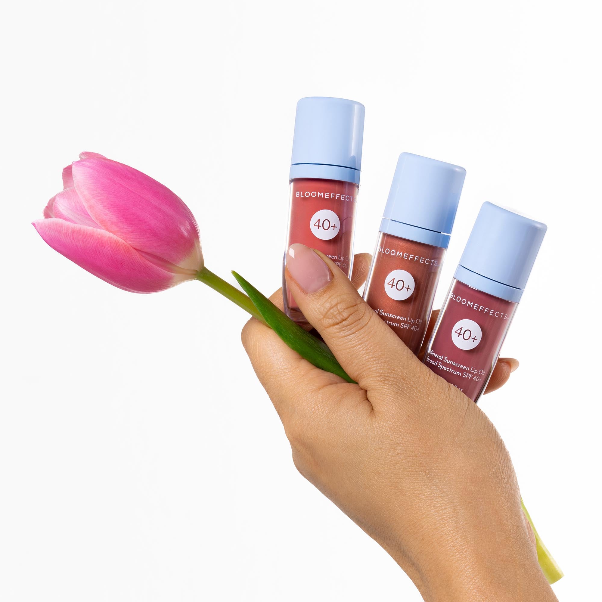 Hand holding three lip oils with a pink tulip against a white background